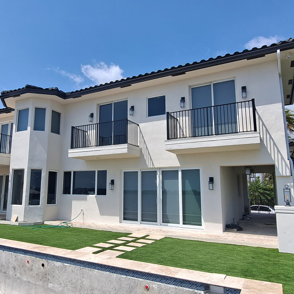 Modern two-story house with a black metal railing balcony, artificial turf yard, and a partially built pool, surrounded by palm trees and a sunny sky 1